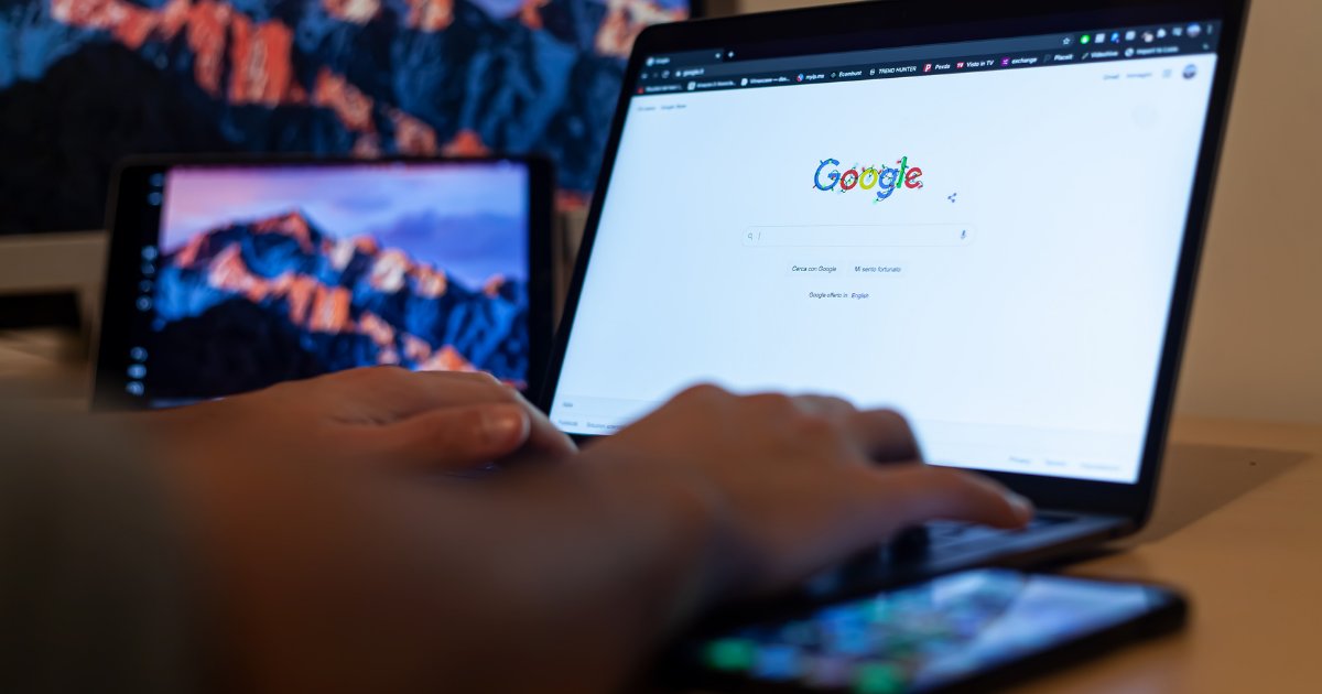 Mão digitando em um notebook com a página inicial do Google aberta, ao lado de um celular e de um monitor